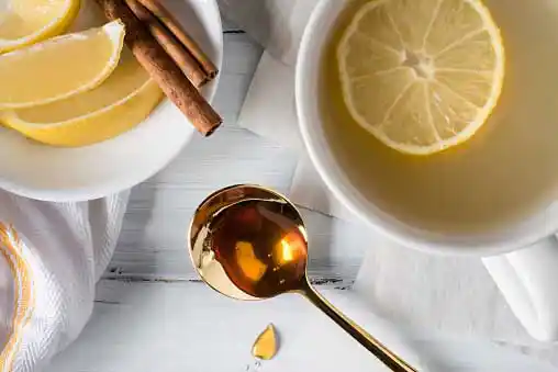slices of cut lemon in a bowl along with a tablespoon of honey and a cup of hot water with lemon piece dipped in it